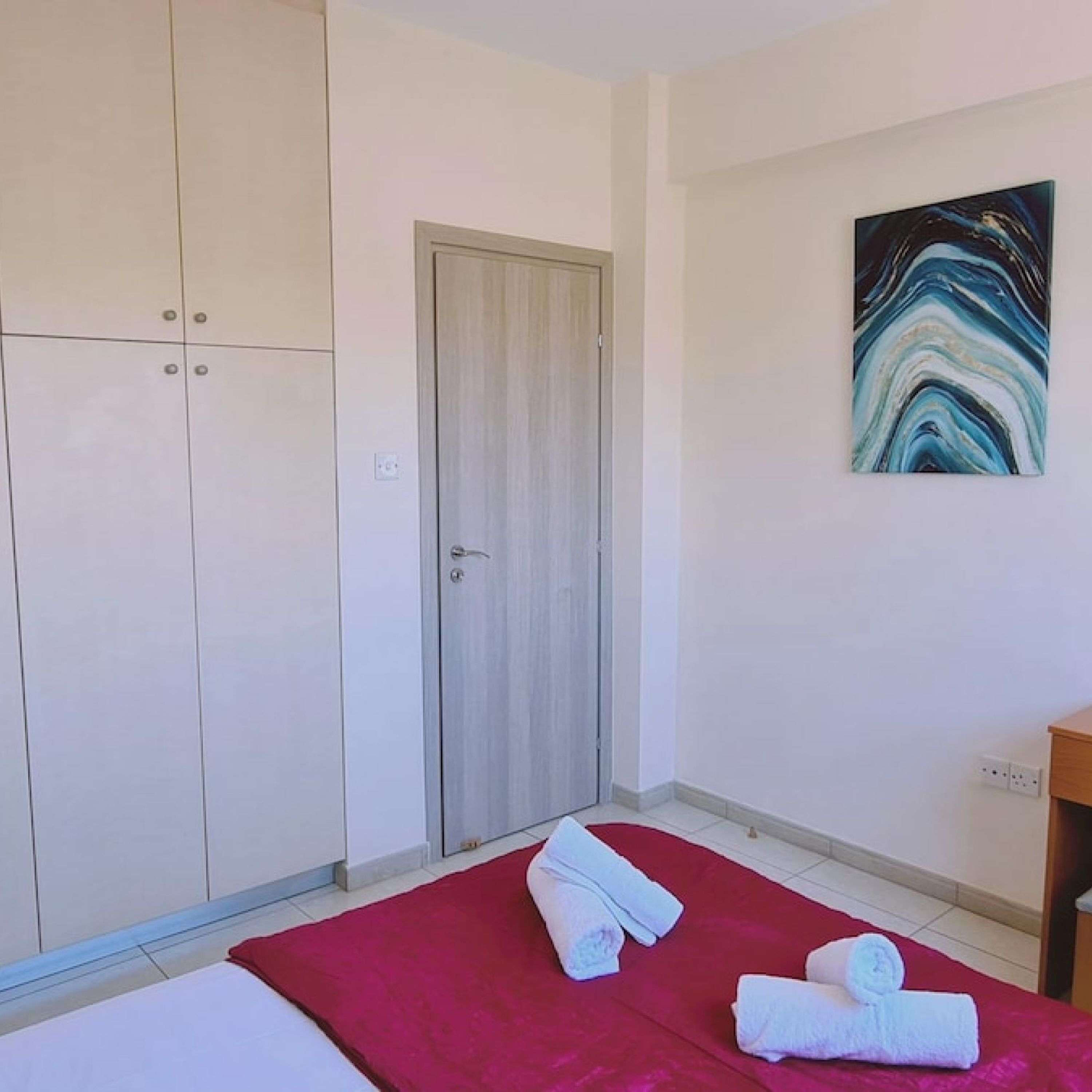 A hotel room with a bed, a red blanket, two towels, a wardrobe, and a painting on the wall.