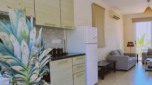A modern kitchen with light wood cabinets, a white refrigerator, and a dining area with a grey sofa and a small table. - 51 PER - Sunny Bay Villa (Paralimni)