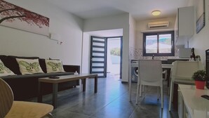 A living room with a sofa, a coffee table, and a TV. There is a kitchen area with a window and an air conditioner. - 14 AN - 114 Nissi Twins Apartment (Ayia Napa)