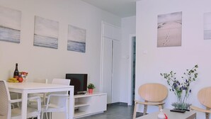 A dining area with a white table, chairs, a television, and artwork on the walls. - 14 AN - 114 Nissi Twins Apartment (Ayia Napa)