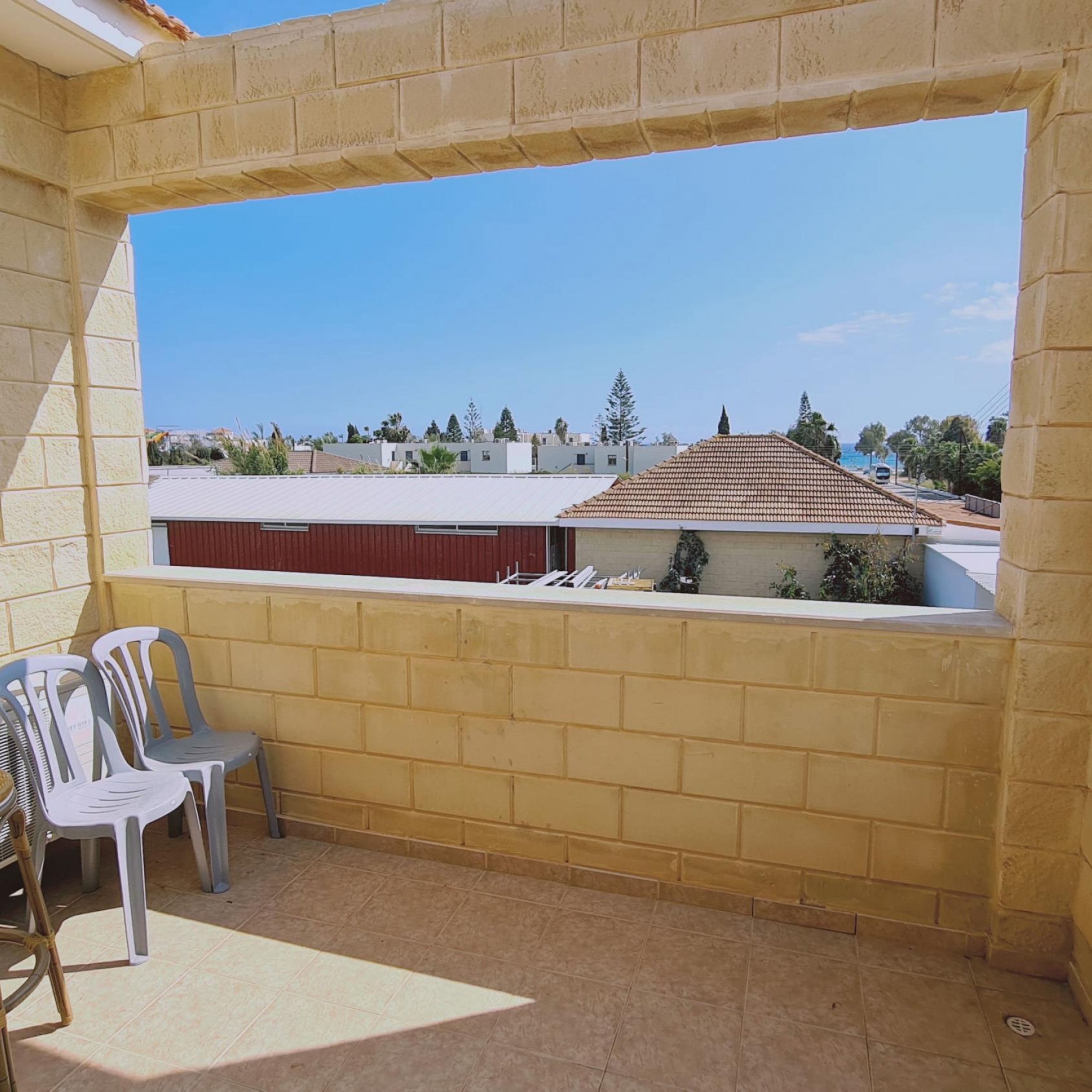 Een balkon met uitzicht op een woonwijk, een rood gebouw en een pannendak. Op het balkon staan twee plastic stoelen en een tafel.