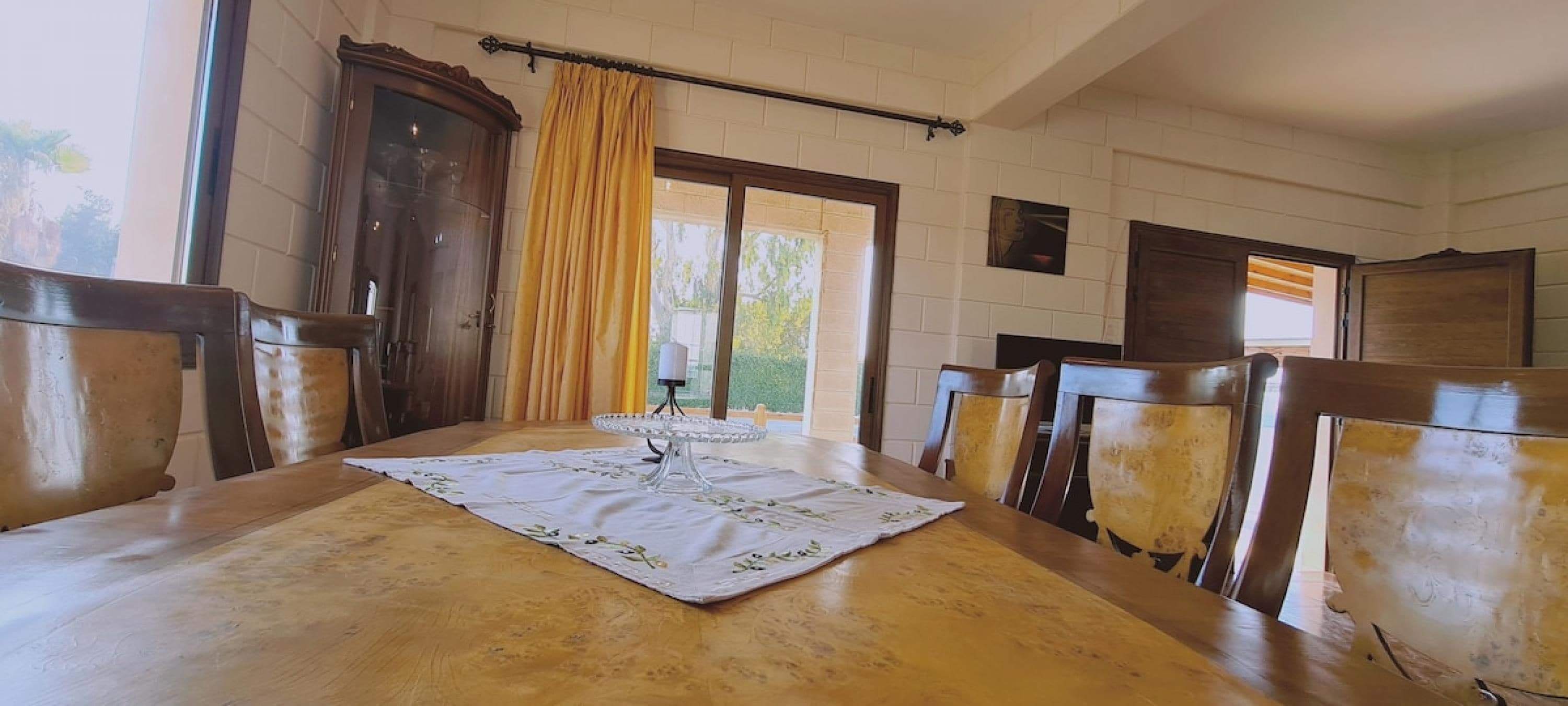 Un comedor con una mesa de madera, sillas, un expositor de vidrio para pasteles y un ventanal con cortinas.