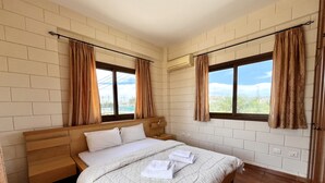 A bedroom with a wooden bed, two windows with curtains, brick walls, and an air conditioning unit. - 25 AN - Pernera House (Ayia Napa)