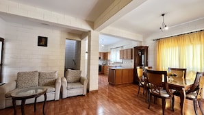 A spacious living area with a sofa, coffee table, and dining table. A kitchen is visible in the background. - 25 AN - Pernera House (Ayia Napa)