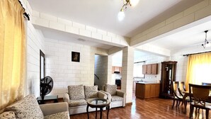A spacious living room with a sofa, armchairs, and a coffee table. A dining area with chairs and a table is visible in the background. - 25 AN - Pernera House (Ayia Napa)