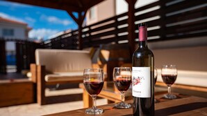 A bottle of wine and three glasses on a wooden table, with a wooden bench and a building in the background. - 08 PER - Villa Anemone (Paralimni)