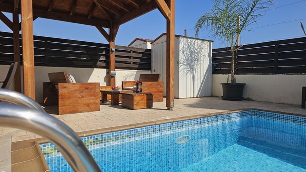 A swimming pool with a tiled edge, wooden furniture, and a potted plant. - 08 PER - Villa Anemone (Paralimni)