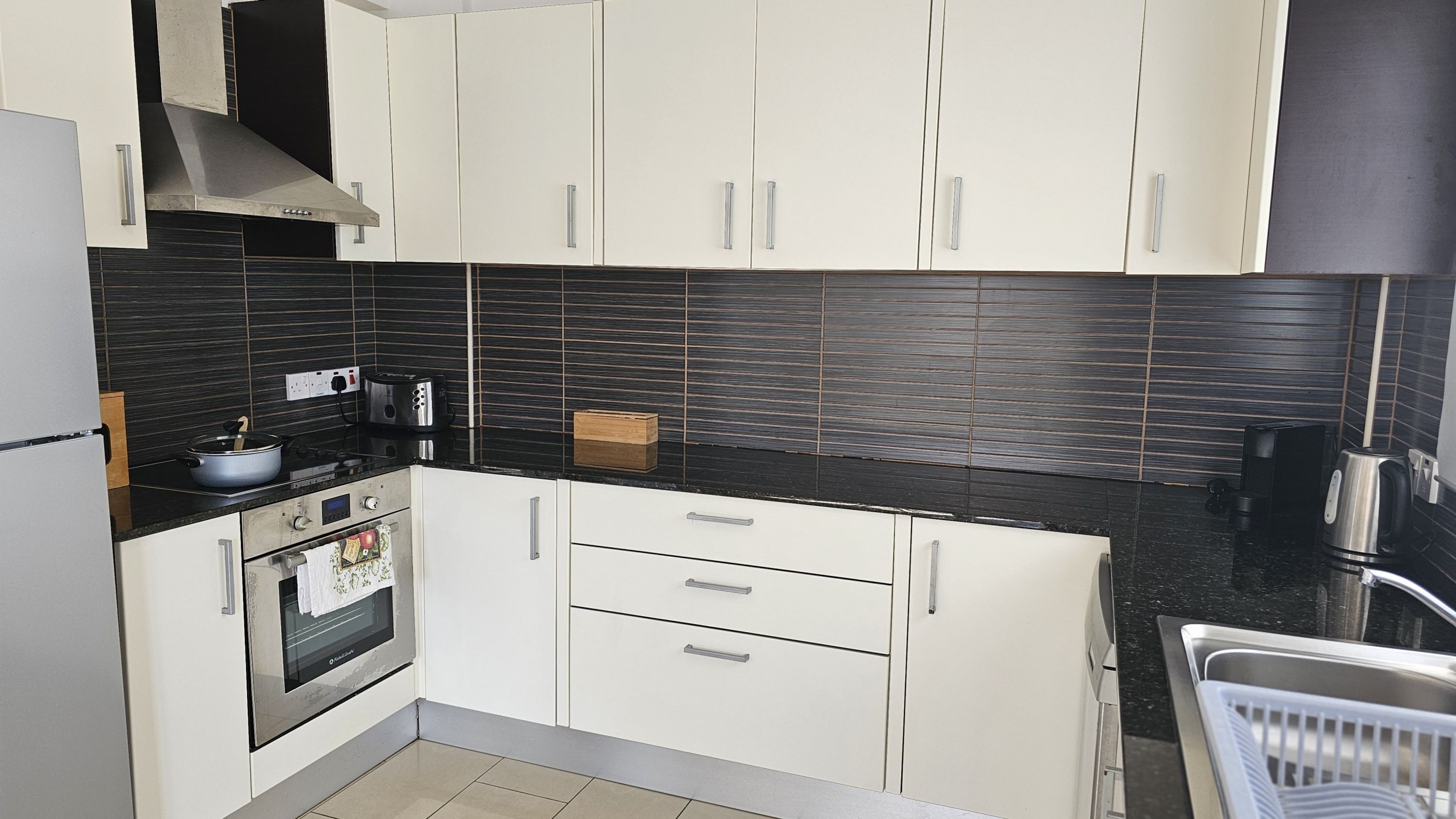 A modern kitchen with white cabinets, a black tiled backsplash, stainless steel appliances, and a built-in oven.