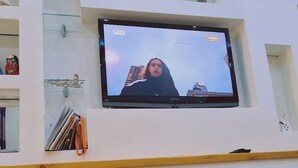 En tv-apparat är monterad på väggen och visar en stadsbild. Under tv:n finns hyllor med böcker och dekorativa föremål. Framför tv:n står en ask med pappersnäsdukar och en liten skål.
