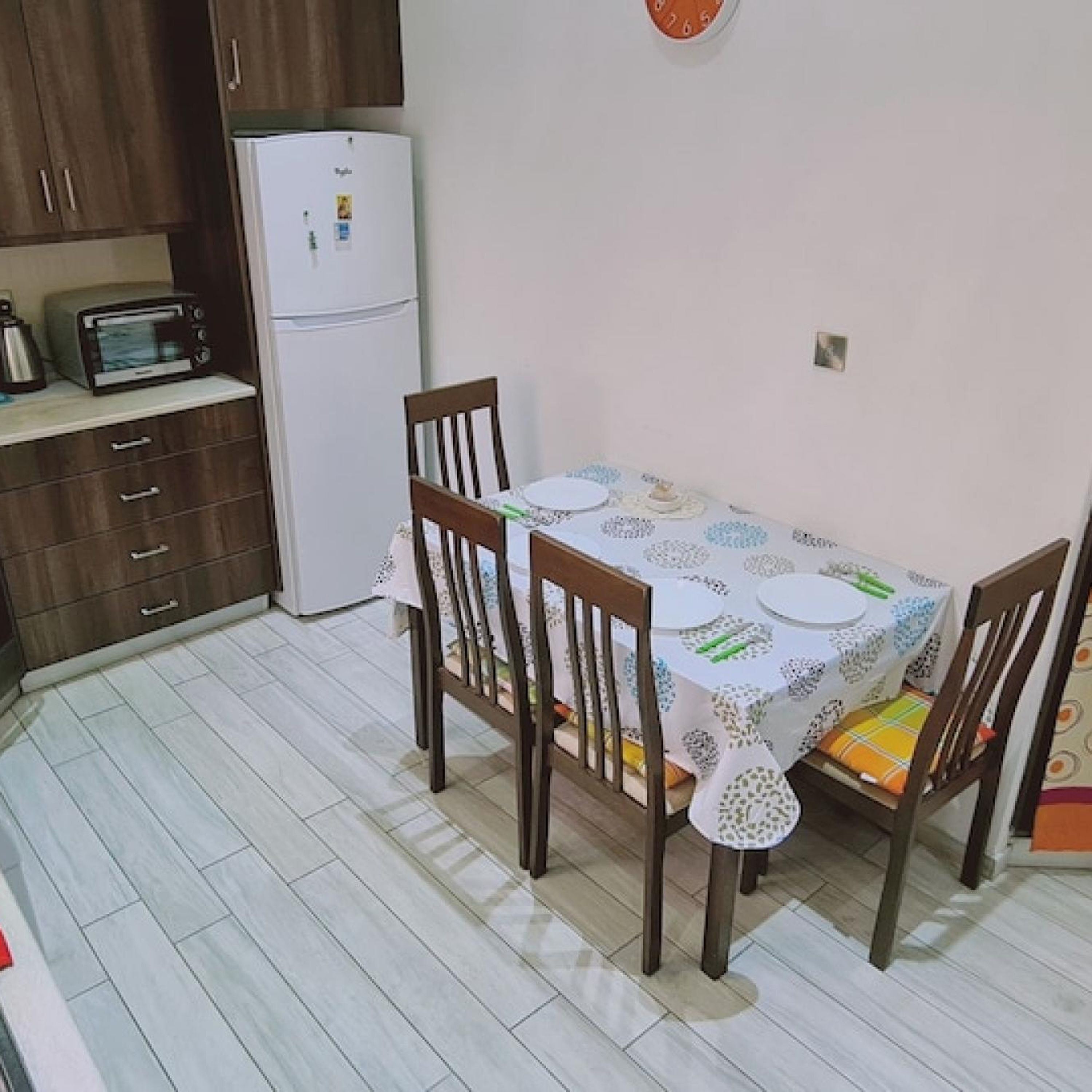 A dining area with a table set for four, surrounded by wooden chairs. A white refrigerator and a toaster are visible in the background.