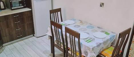 A dining area with a table set for four, surrounded by wooden chairs. A white refrigerator and a toaster are visible in the background.