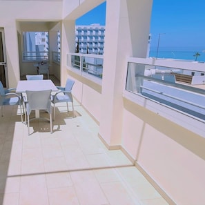 A balcony with a table and chairs, a view of the sea, and a building in the background. - 40 PRO 1 - Sunrise Beach Apt 1 (Paralimni)