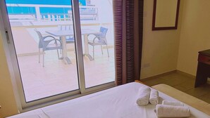 Habitación de hotel con cama, ventana con vistas al mar y balcón con sillas y mesas.