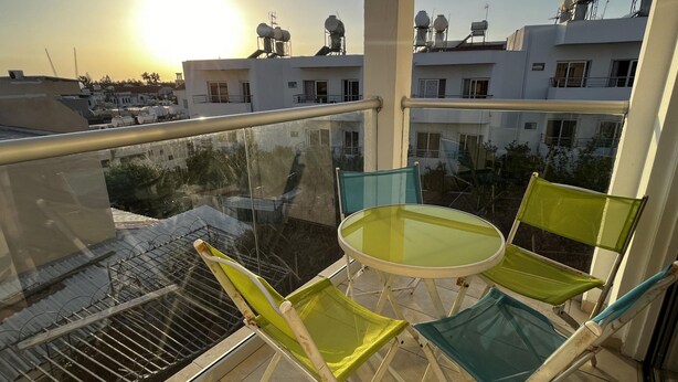 A balcony with a glass table and chairs, overlooking a residential area with buildings and a clear sky.