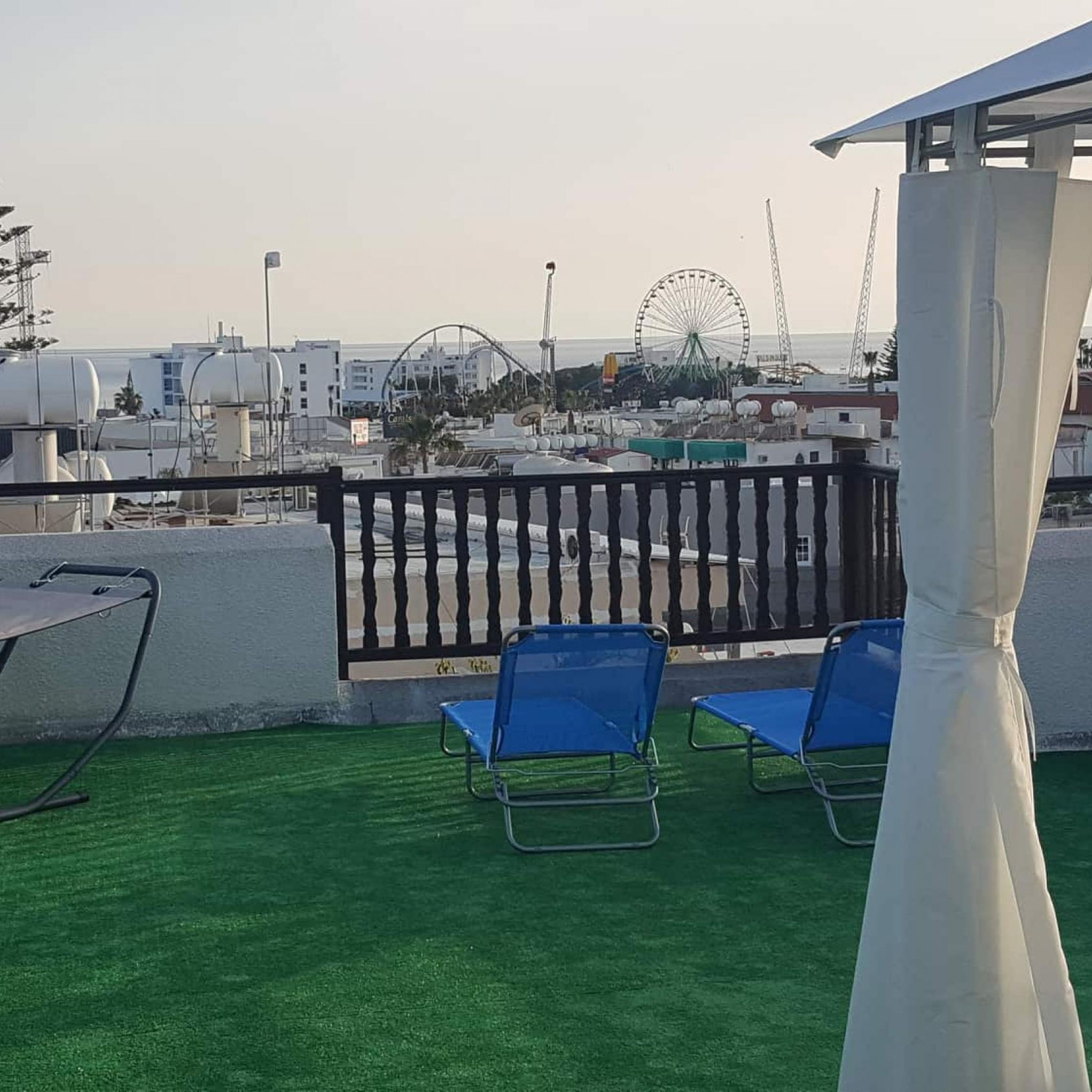 Una terraza en la azotea con césped artificial, dos sillones azules, un toldo blanco y vistas a una noria y edificios industriales al fondo.