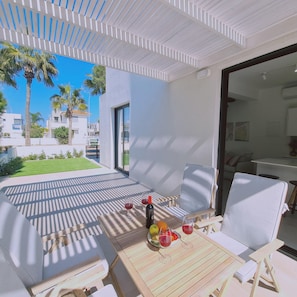A modern outdoor patio with a wooden table set for two, a sofa, and a chair. There are two wine glasses and a bottle on the table. - Stay Sunshine Aura in Protaras (Paralimni)