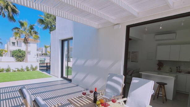 A modern outdoor dining area with a table set for two, a kitchen visible through an open door, and a view of a residential area with palm trees. - Stay Sunshine Aura in Protaras (Paralimni)
