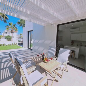 Een modern terras met eettafel gedekt voor twee personen, met een keuken zichtbaar door een open deur, en uitzicht op een woonwijk met palmbomen.