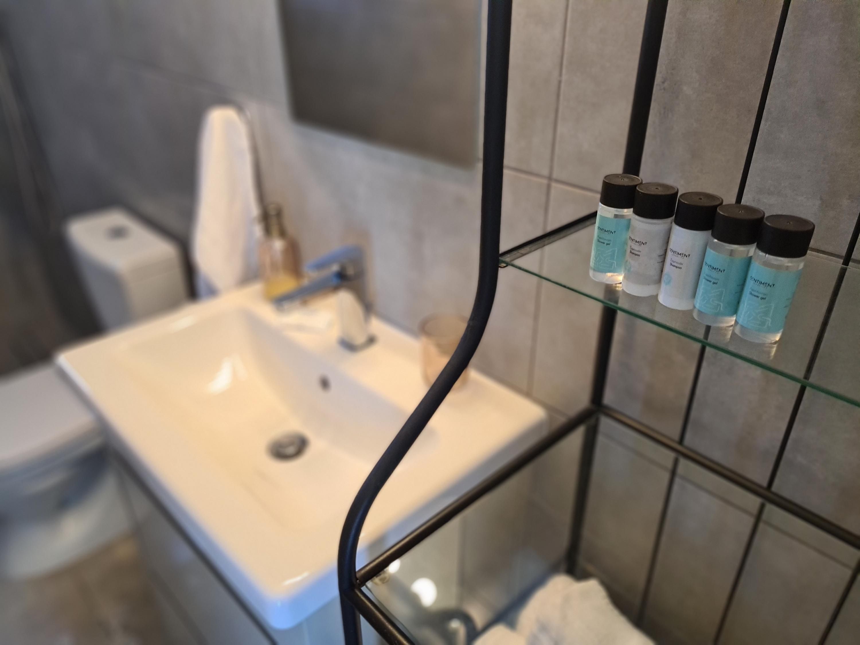Un baño con un lavamanos blanco, un estante de vidrio con artículos de aseo y una toalla colgada en la pared.