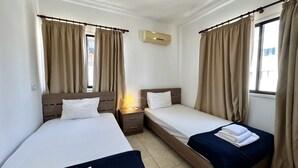 Ein Hotelzimmer mit zwei Einzelbetten, einem Nachttisch mit Lampe, einer Klimaanlage und beigen Vorhängen.