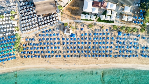 Ein Luftbild eines Strandresorts mit Reihen blauer Sonnenschirme, einem Sandstrand und klarem türkisblauem Wasser.