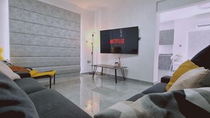 Een moderne woonkamer met een grote flatscreen televisie aan de muur, een tafeltje met een fles en een bank met kussens.
