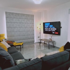 Een moderne woonkamer met een grote flatscreen televisie aan de wand, een grijze bank met gele kussens, een klein houten bijzettafeltje en een beton accentmuur.