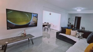 Een moderne woonkamer met een flatscreen televisie, een houten bankje, een bank met kussens, en een eethoek op de achtergrond.