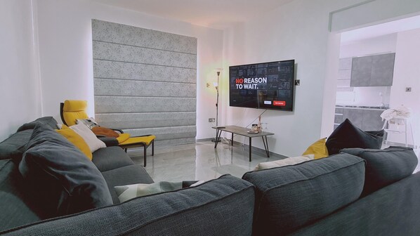 Een moderne woonkamer met een grijze bank, gele kussens, een flatscreen televisie en een keuken op de achtergrond.