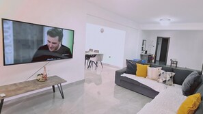 Een moderne woonkamer met een flatscreen televisie, een houten salontafel, een grijze bank met kussens, en een eethoek op de achtergrond.