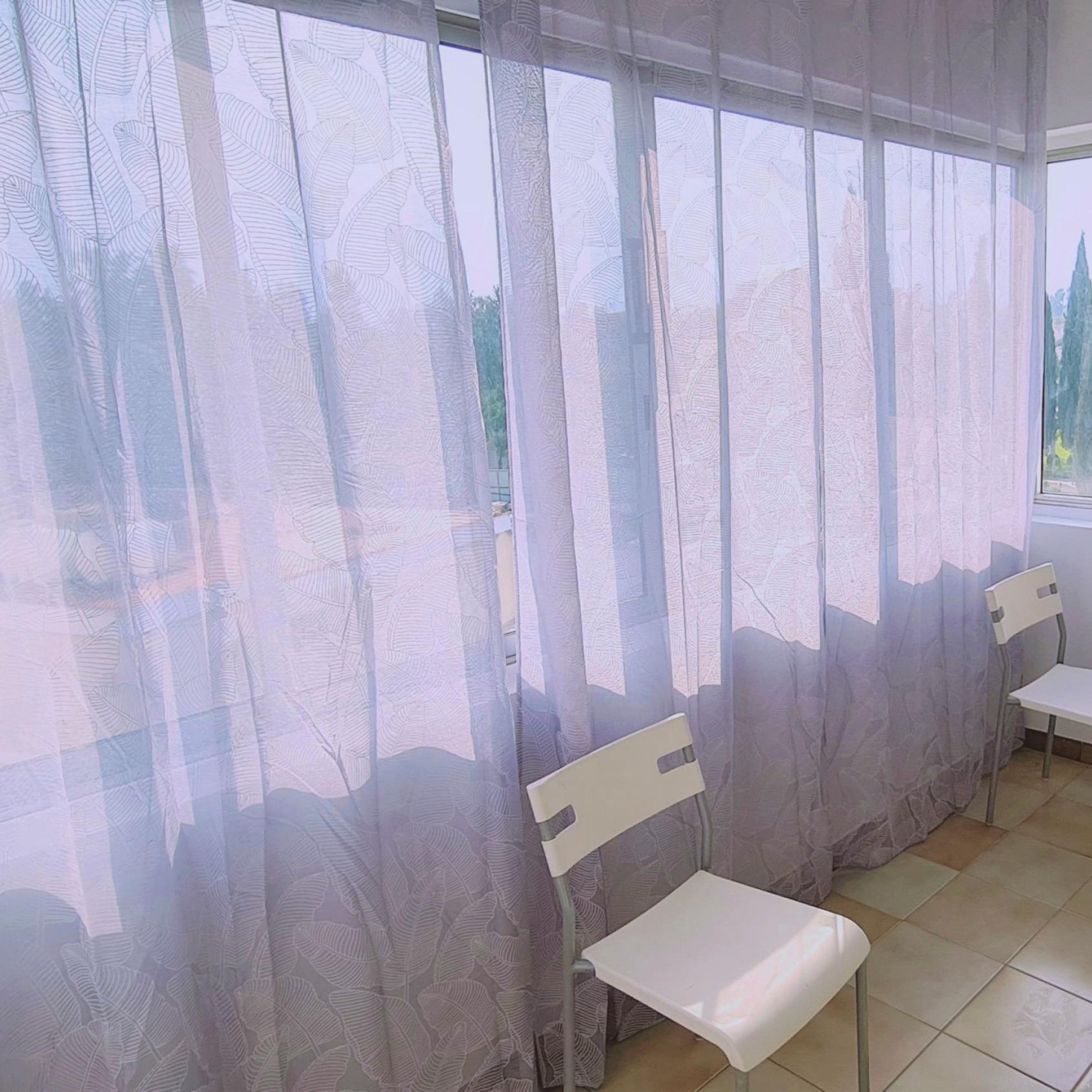 Una habitación con un ventanal grande cubierto por cortinas translúcidas, dos sillas blancas y un suelo de baldosas.