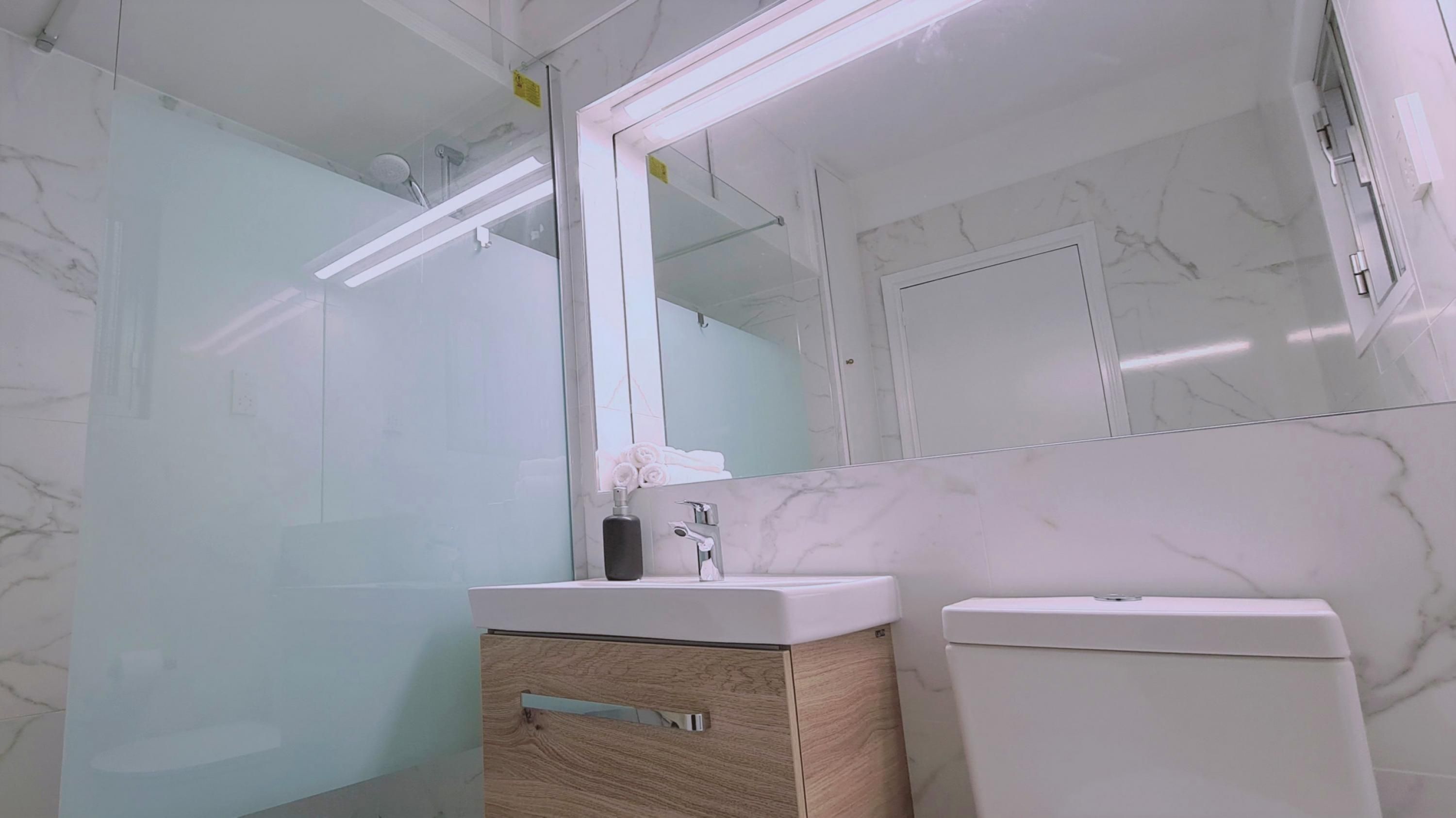 A modern bathroom with a glass shower enclosure, a marble-tiled wall, a wooden vanity with a mirror, and a toilet.