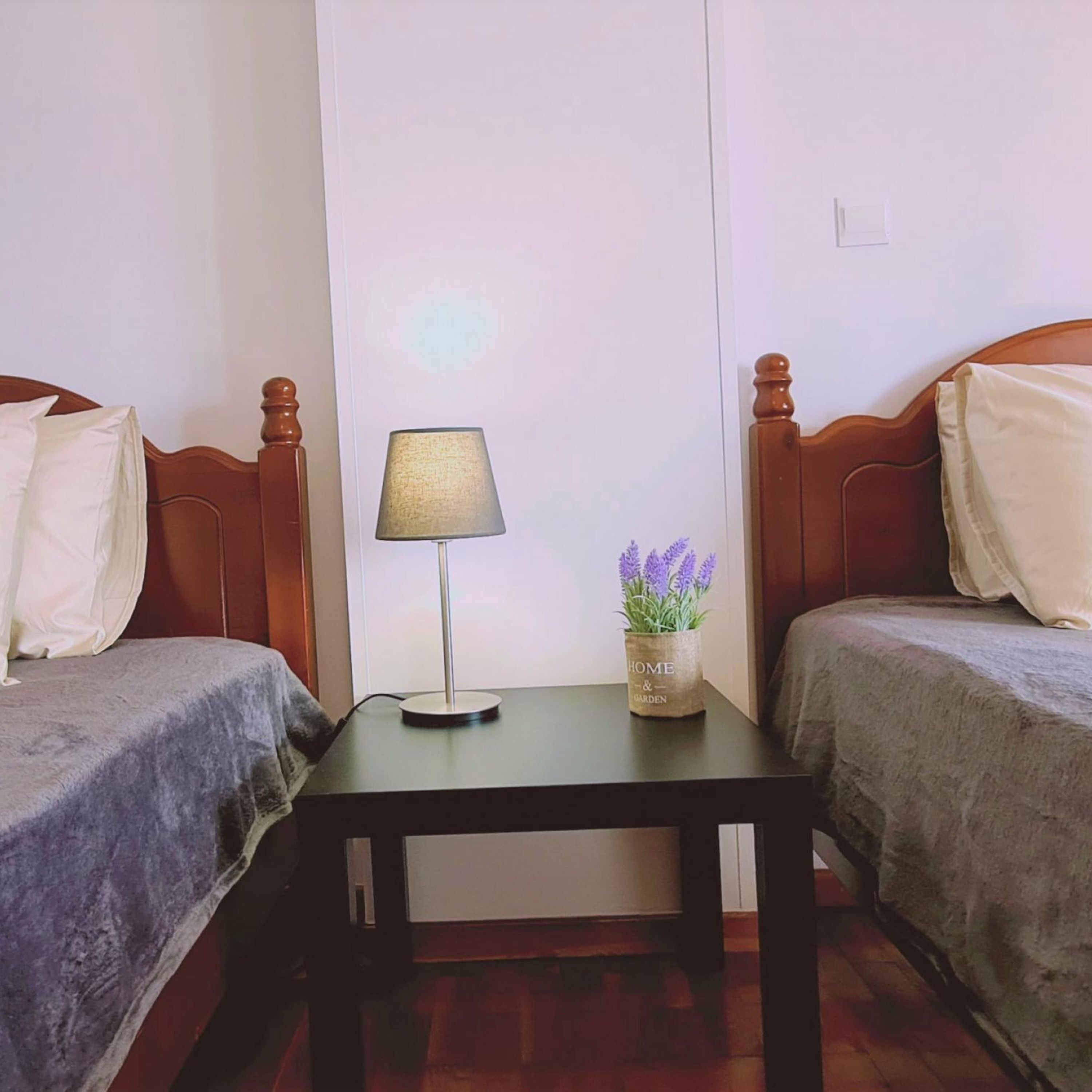 A room with two wooden beds, a small table with a lamp, and a vase with flowers.