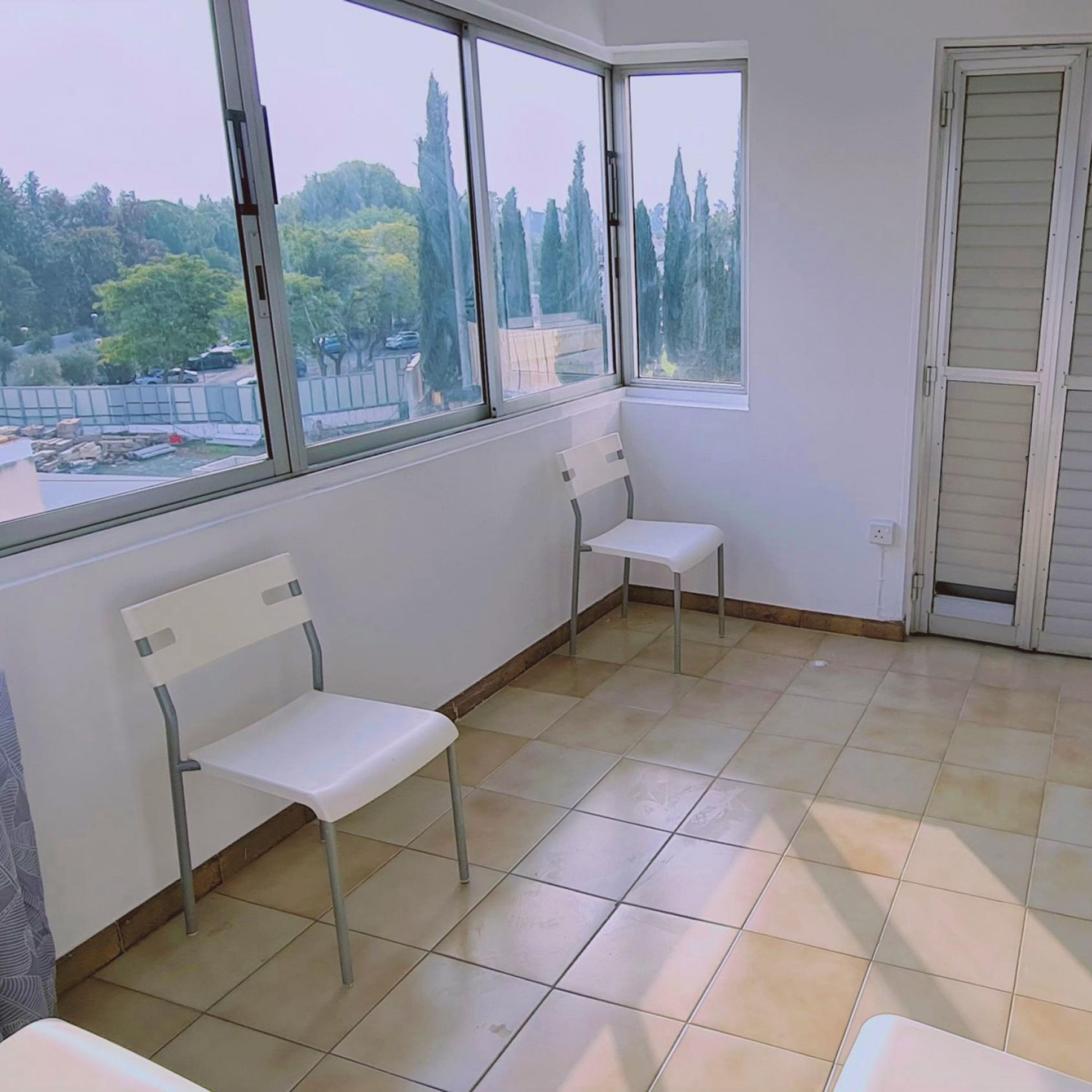 A room with two white chairs, a tiled floor, and large windows offering a view of trees and a fence.