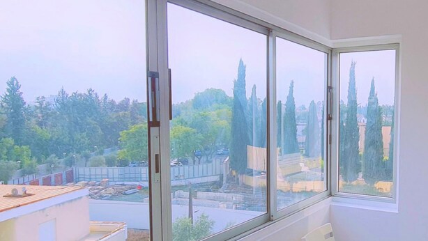 Ein Zimmer mit großem Fenster und Blick auf Bäume sowie eine Baustelle. Ein weißer Stuhl steht in der Nähe des Fensters.
