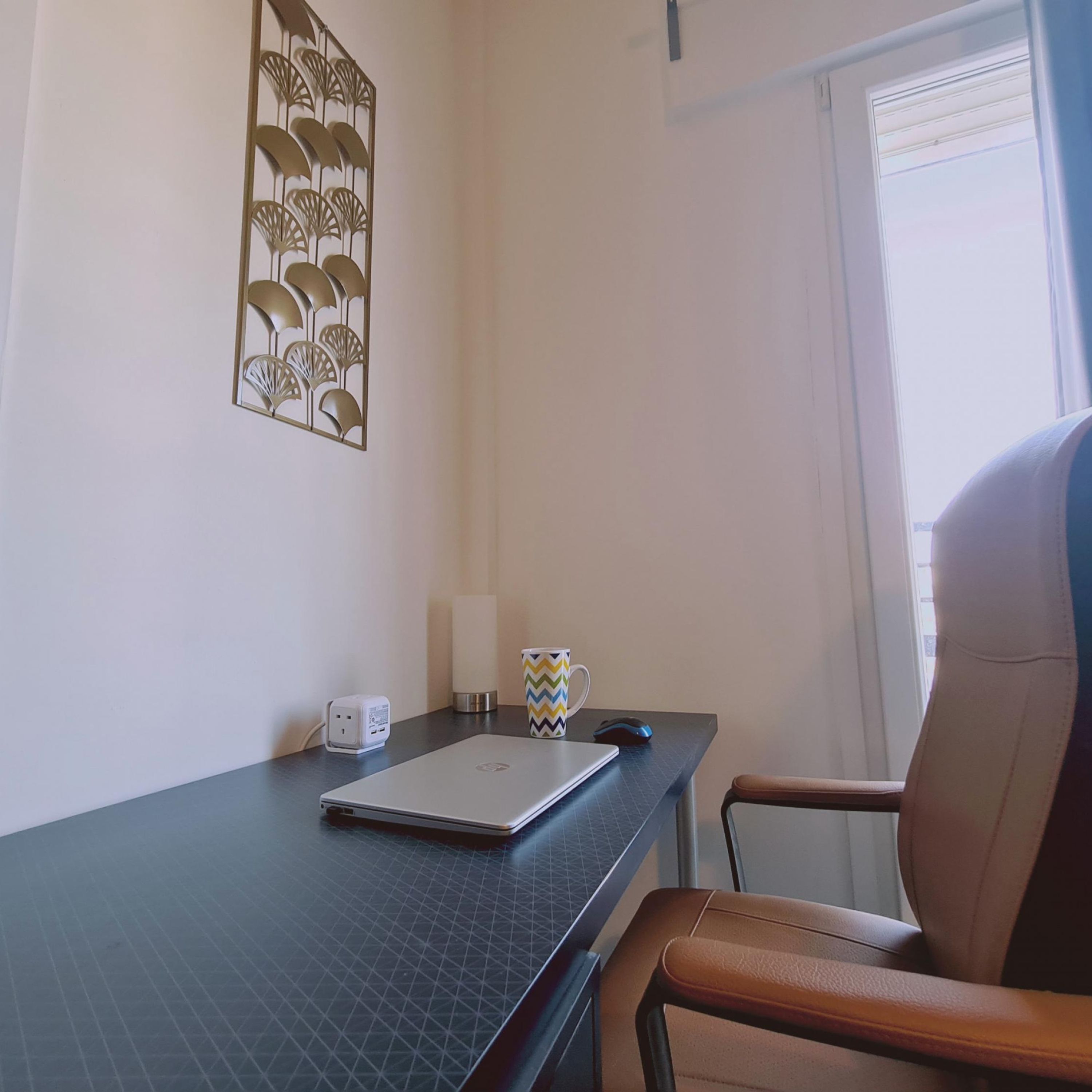 A room with a desk, a laptop, a chair, a decorative wall piece, a cup, and a mouse.