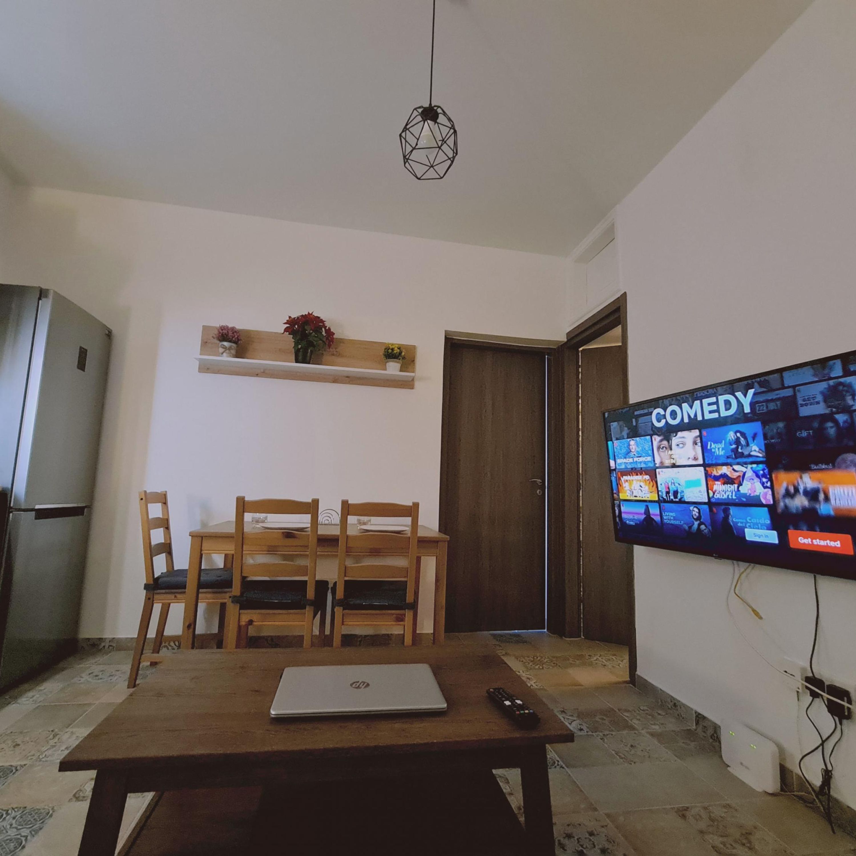 A dining area with a wooden table, chairs, a laptop, and a television displaying a comedy channel.