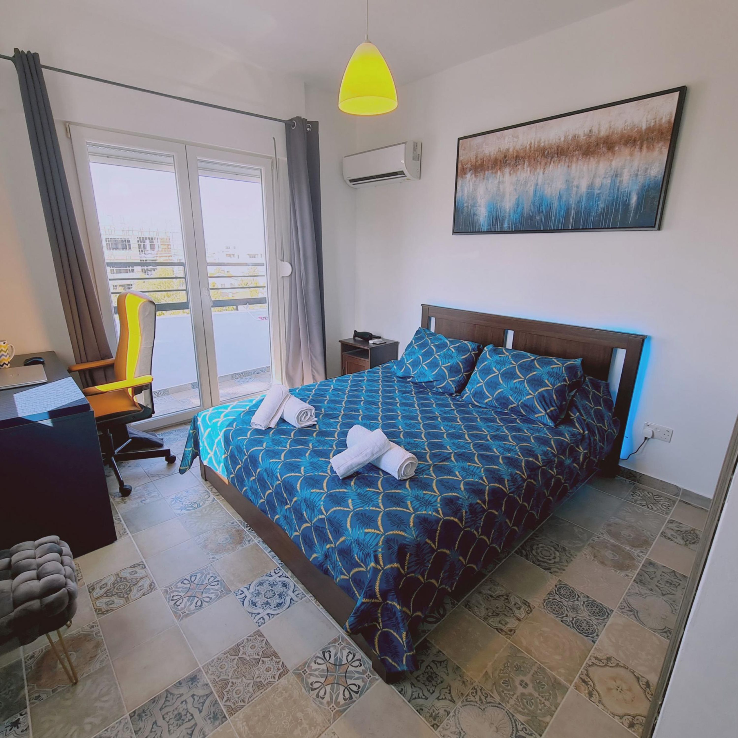 A hotel room with a bed, a desk, a chair, a desk lamp, an air conditioner, and a window with curtains.