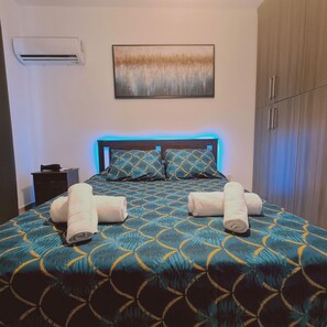 A neatly made bed with a patterned bedspread, two rolled towels, a nightstand, a wall-mounted air conditioner, and a piece of abstract art above the bed. - 18 NIC - Stay Kallipoleos Panorama (Nicosia)