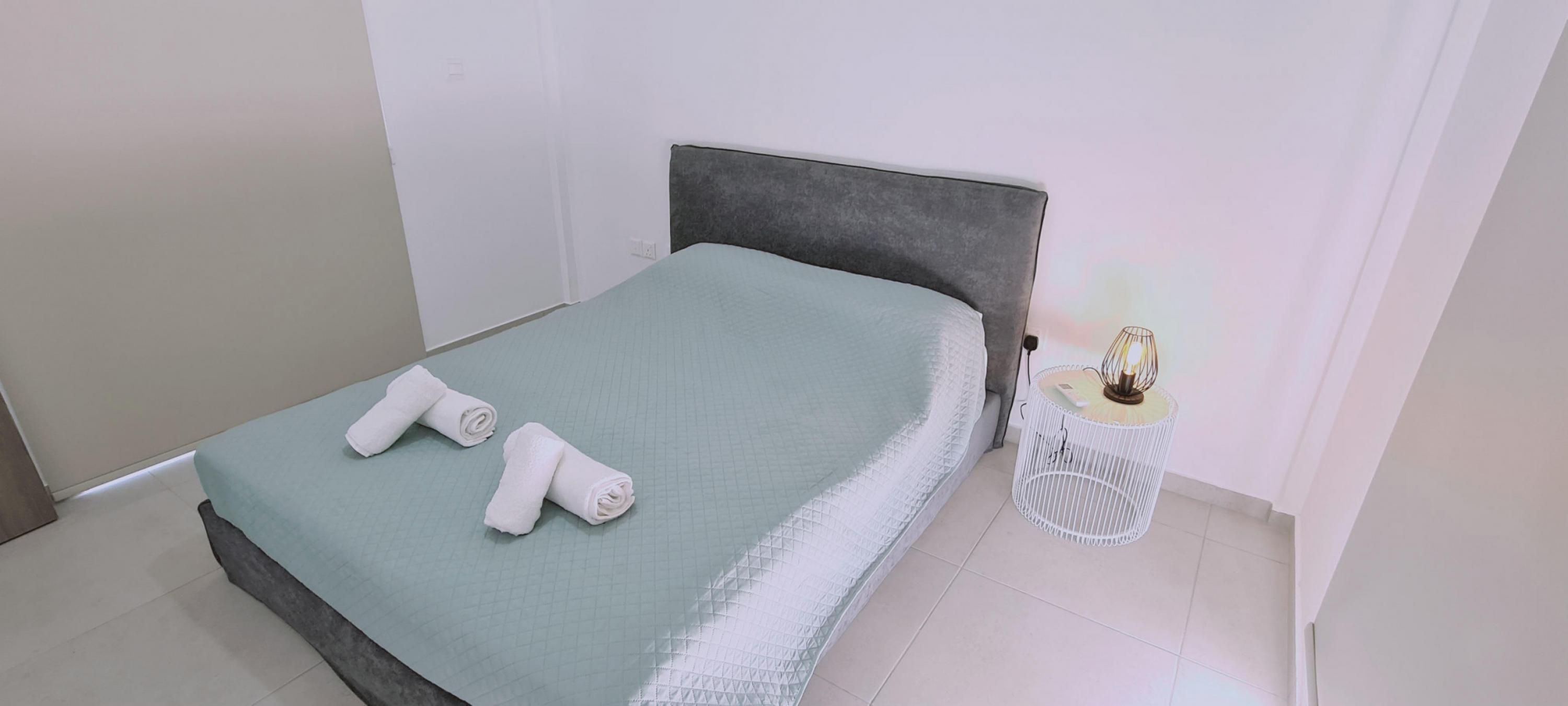 A neatly made bed with a grey headboard, a white bedside table with a lamp, and a small white basket.