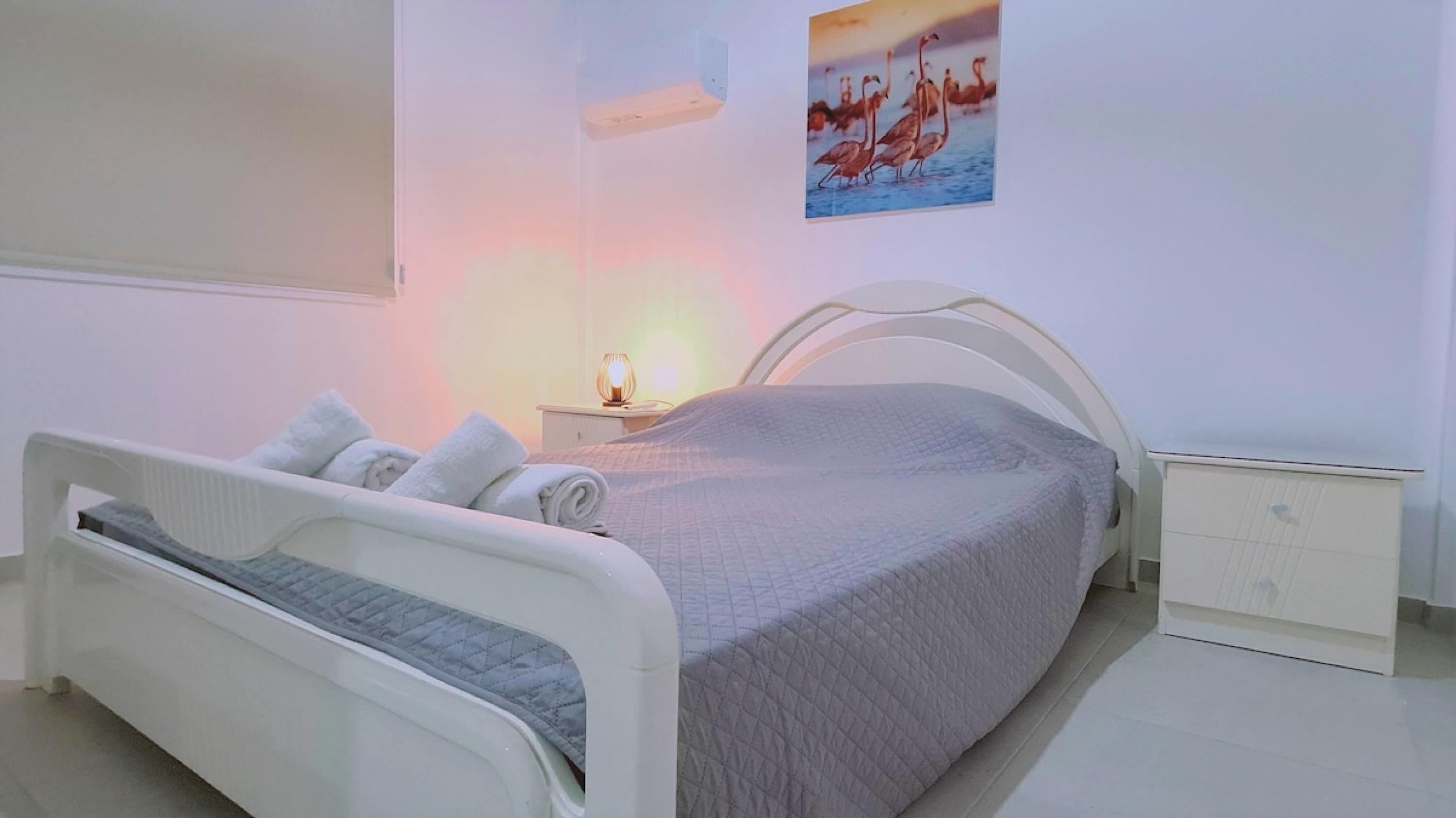 Een keurig opgemaakt bed met kussens en een gewatteerd plaid, een nachtkastje met een lamp, en een aan de muur hangend schilderij van flamingo's.