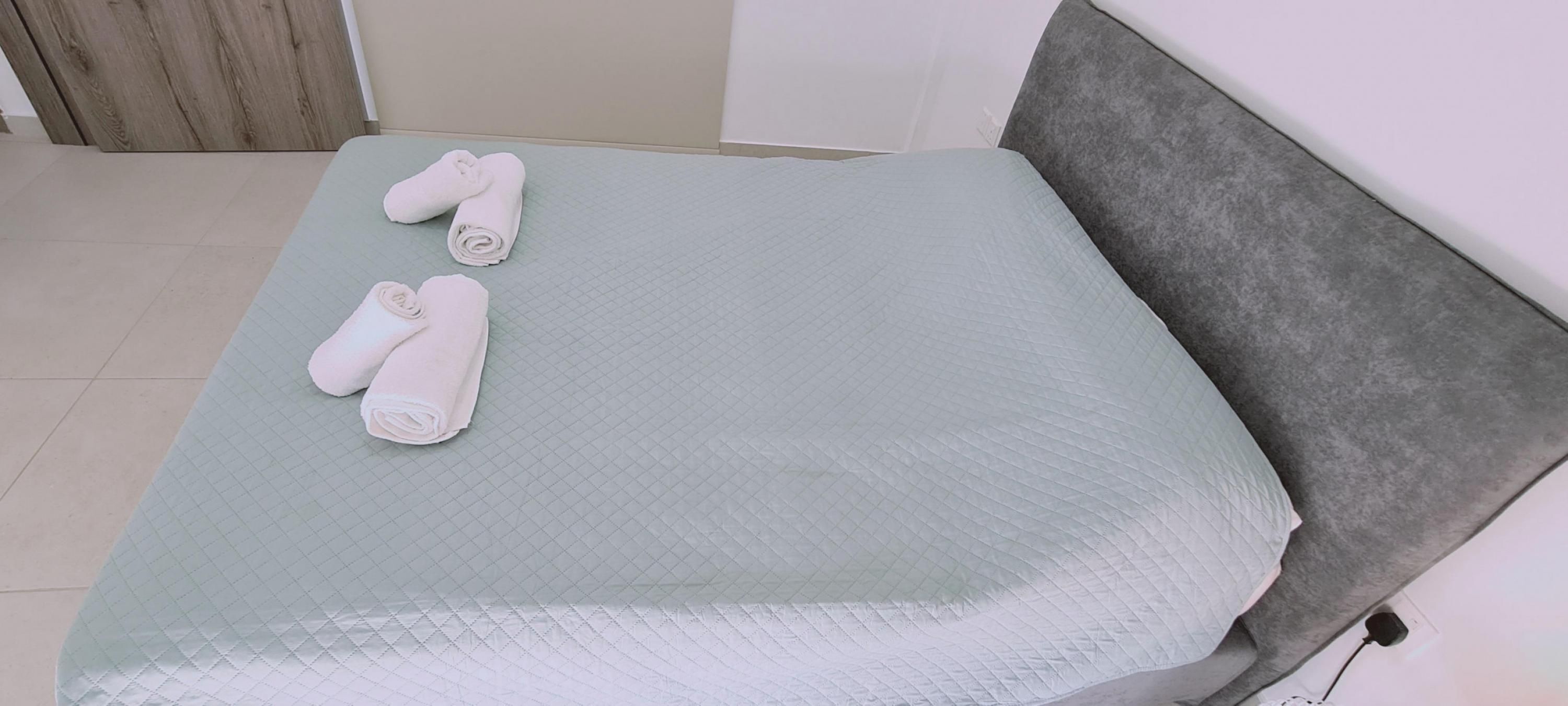 Una cama con colchón acolchado, cabecera gris y dos toallas enrolladas sobre la cama.