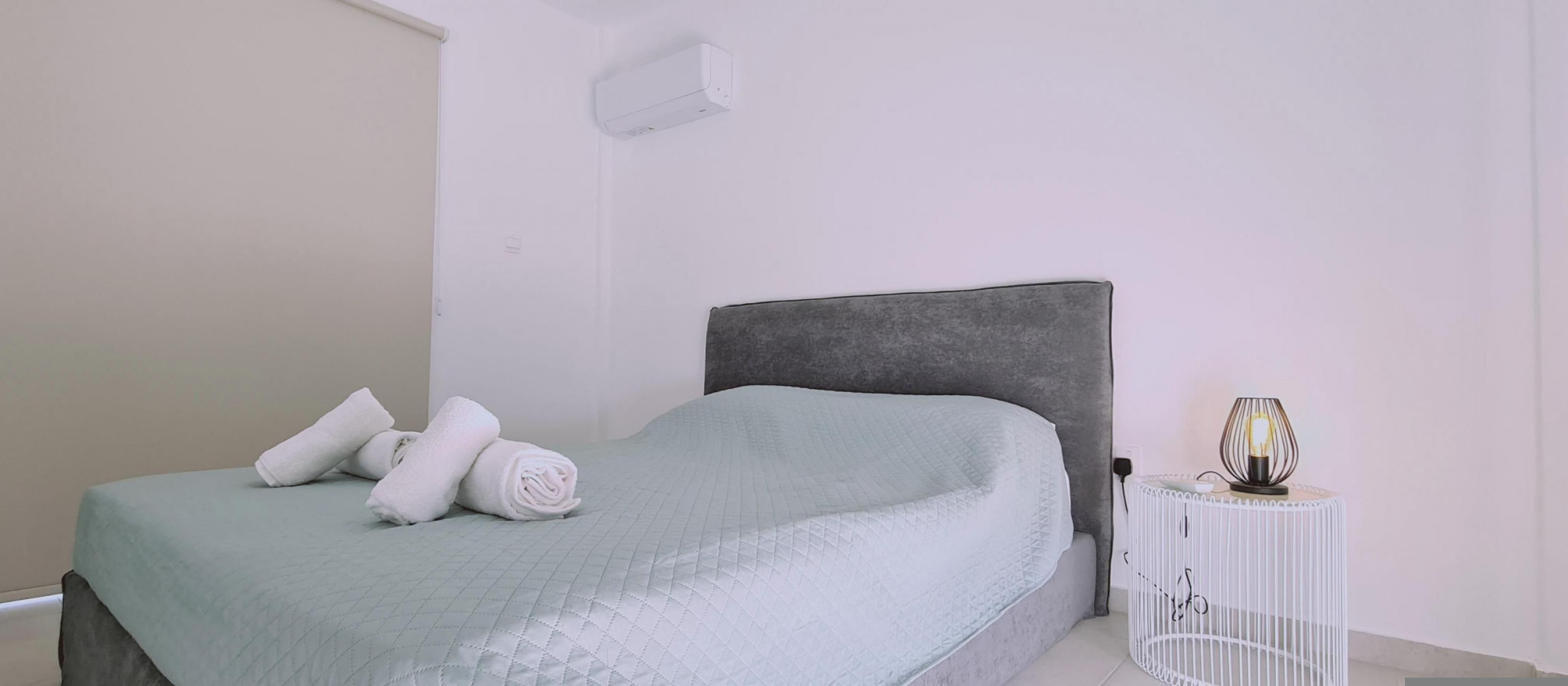 A bed with a quilted cover, a grey headboard, and rolled white towels on it.