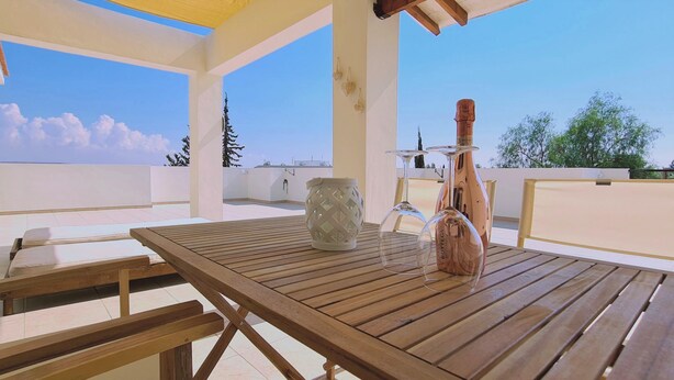 A wooden table with a bottle and a decorative vase on a balcony with a clear view of the sky and trees. - 61 VOR - Sunny Palms (Oroklini)