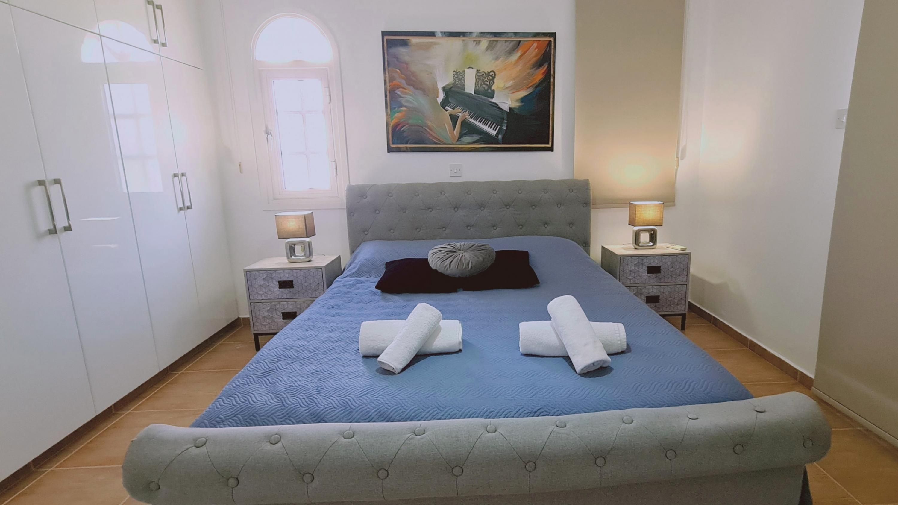 A bedroom with a large bed, two bedside tables with lamps, a painting on the wall, and a wardrobe.