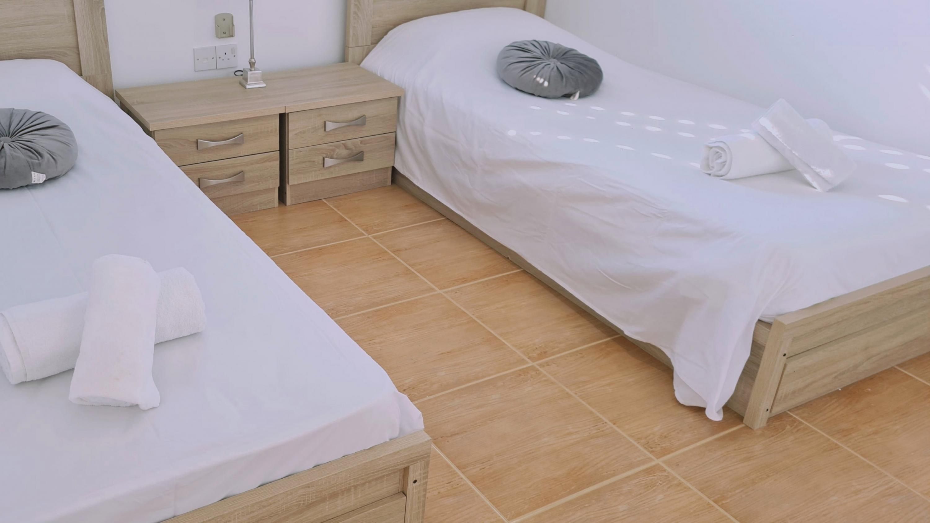 Une chambre d’hôtel avec deux lits simples, une table de chevet en bois avec une lampe et une petite commode.