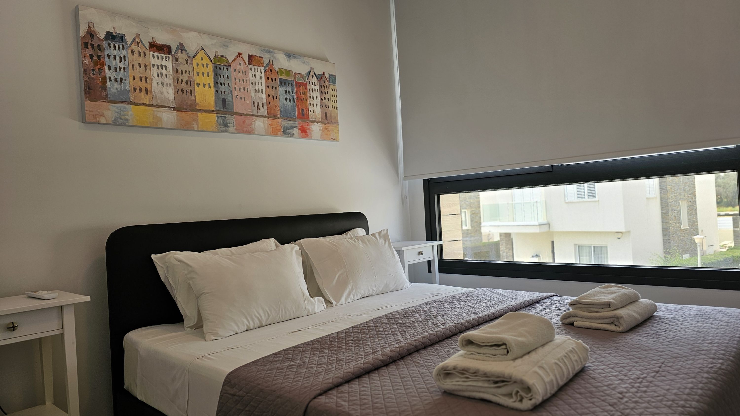 A neatly made bed with pillows and a quilt, a bedside table with a lamp, and a window with a view of buildings.