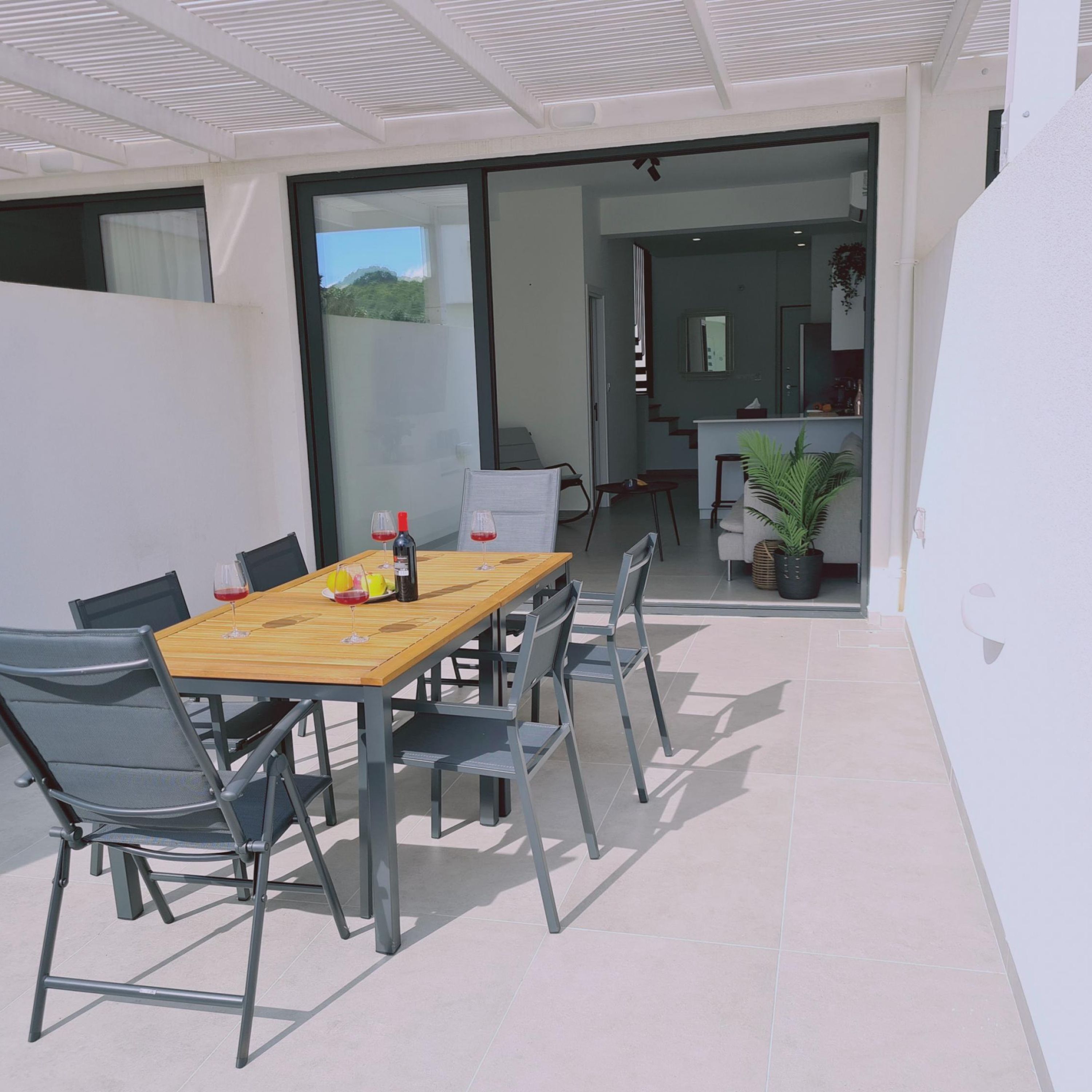 Un espacio exterior moderno para comer con una mesa de madera y sillas de metal, una puerta de vidrio que da a una cocina y una planta en maceta.