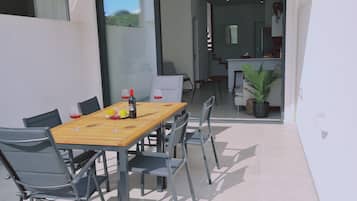 Un espacio exterior moderno para comer con una mesa de madera y sillas de metal, una puerta de vidrio que da a una cocina y una planta en maceta.