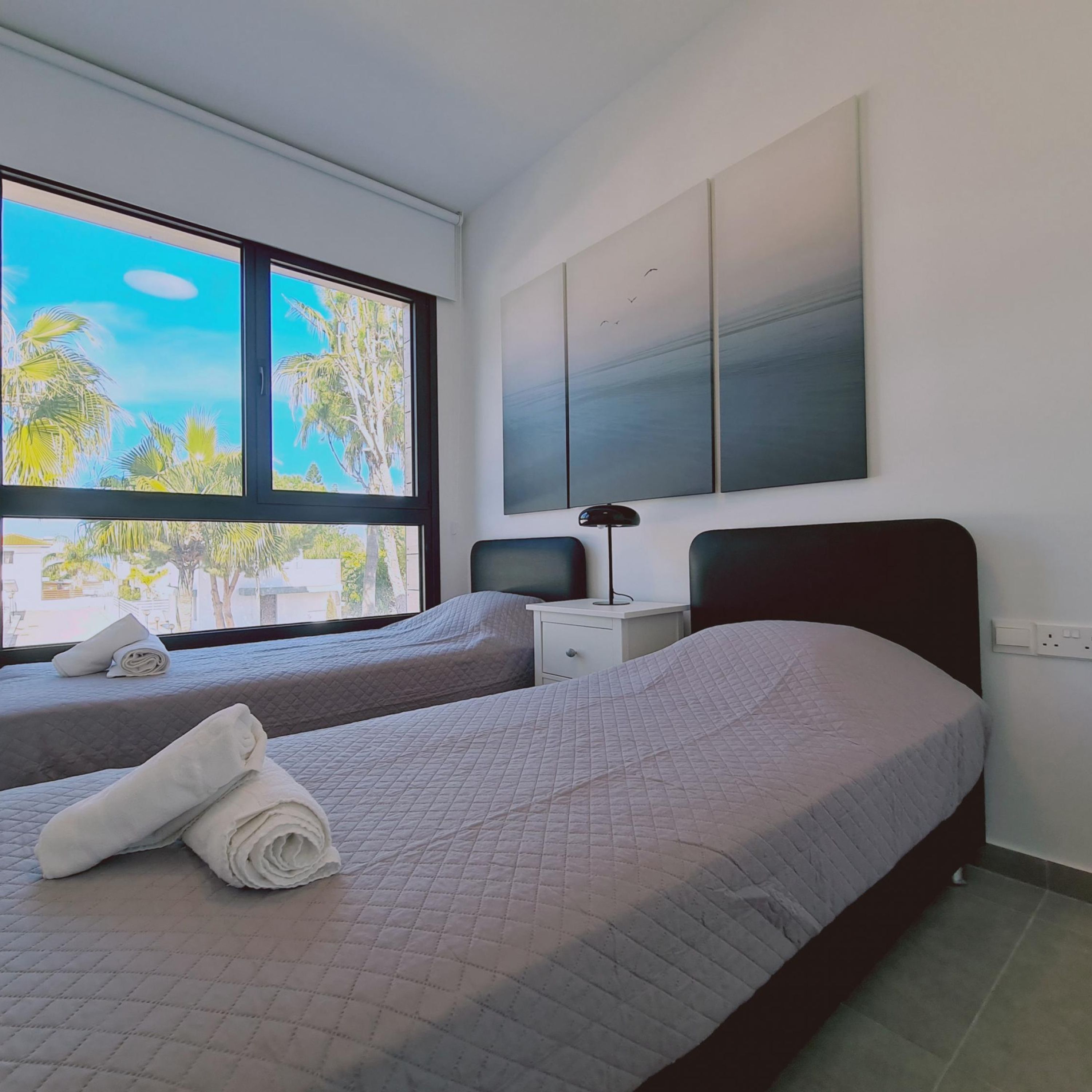 A hotel room with two beds, a nightstand, a lamp, and a large window with a view of palm trees.